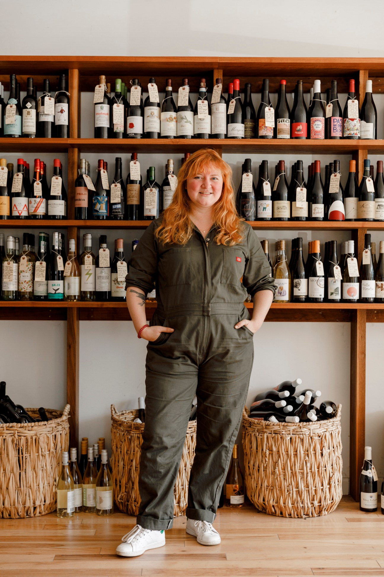 Woman with red hair standing in front of wooden wine shelves wearing olive-green jumpsuit.