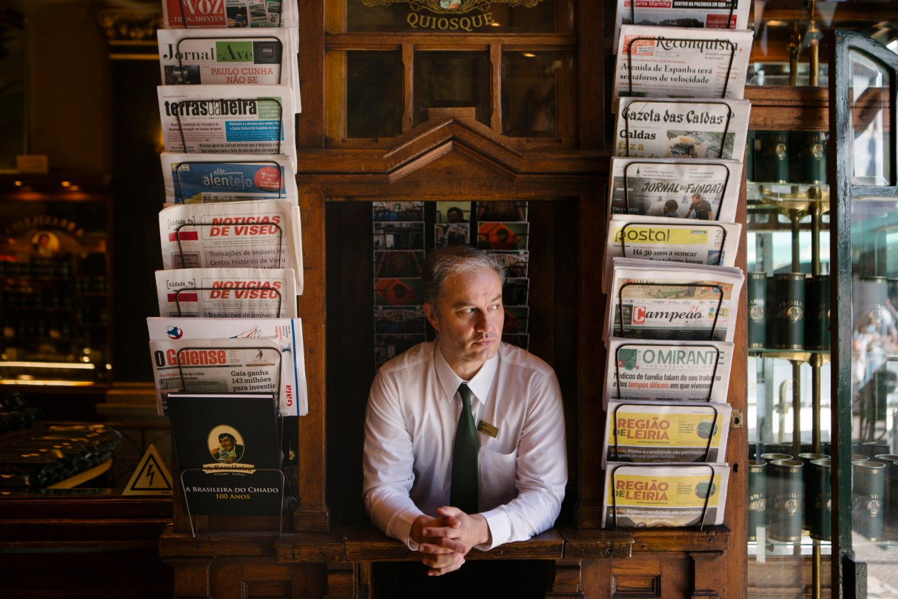 Man in white shirt and green tie standing at A Brasileira café newsstand surrounded by Portuguese newspapers and magazines.
