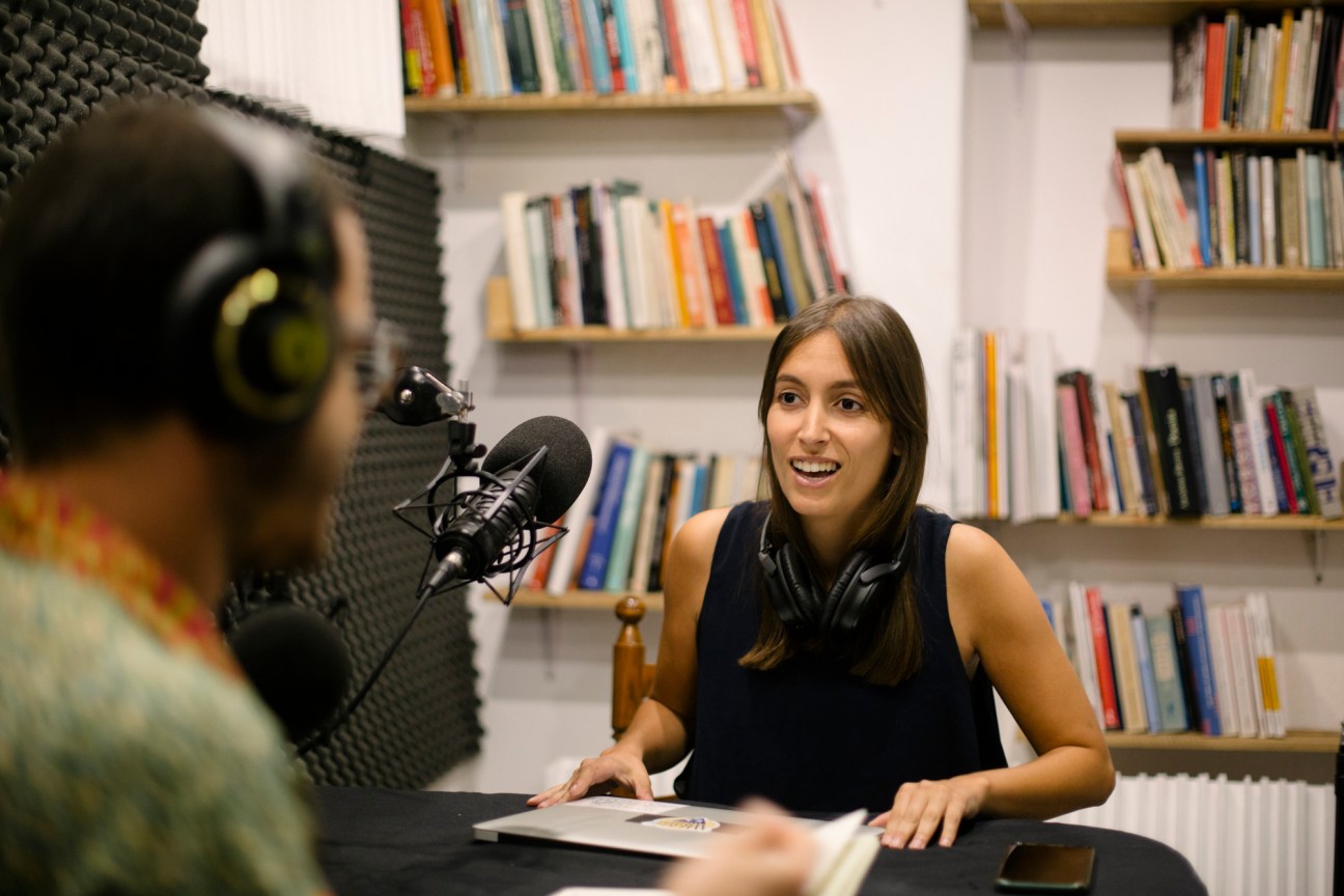 Woman speaking into a microphone at a desk with bookshelves behind her in a studio setting.