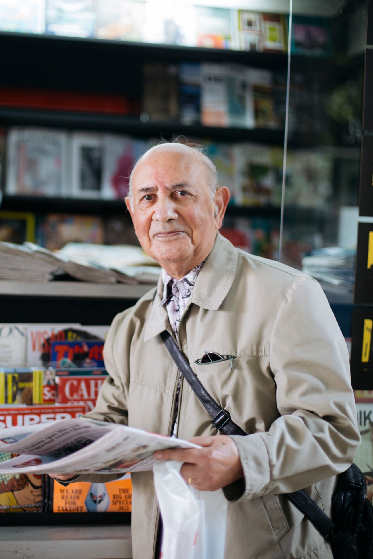 An older man in a beige jacket holds a newspaper while standing in front of shelves of publications.