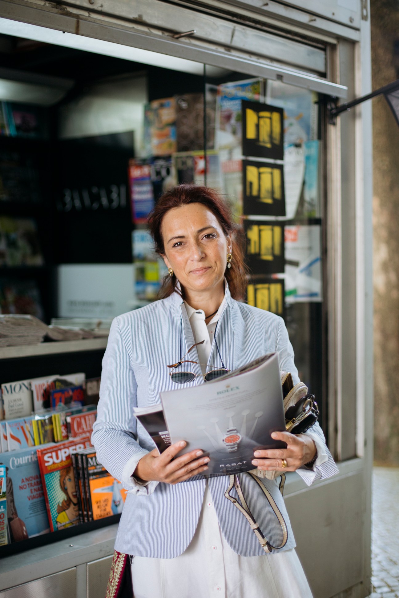 Woman in striped blazer holding newspaper and magazines outside storefront with yellow signage
