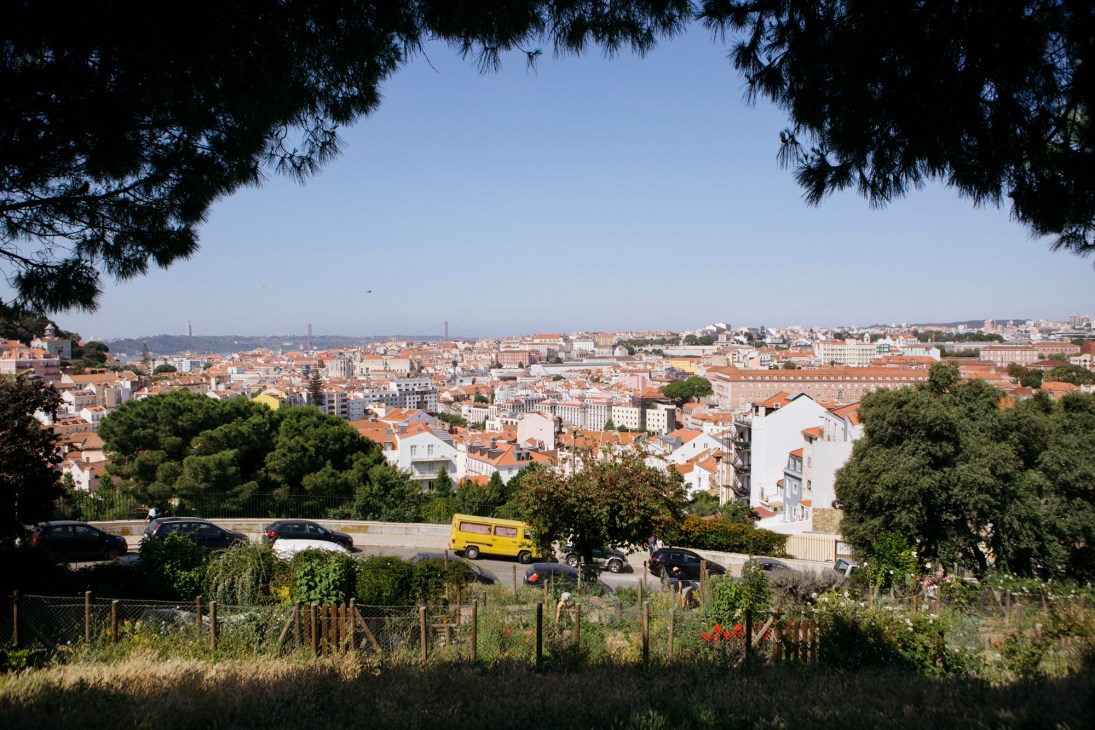 Cityscape of Lisbon with terracotta-roofed buildings, a yellow van, and suspension bridge visible in the distance under blue sky.