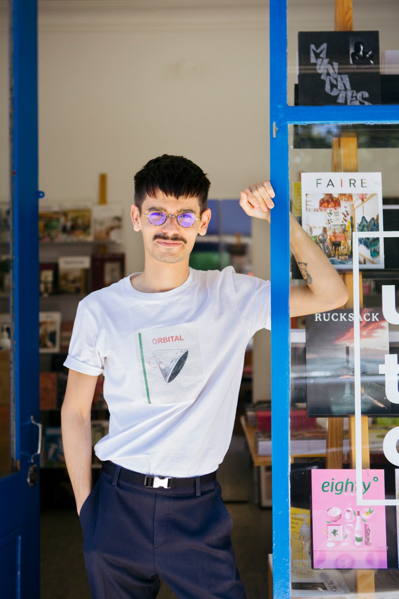 Young man in purple glasses and white t-shirt posing inside a magazine shop with blue framing.