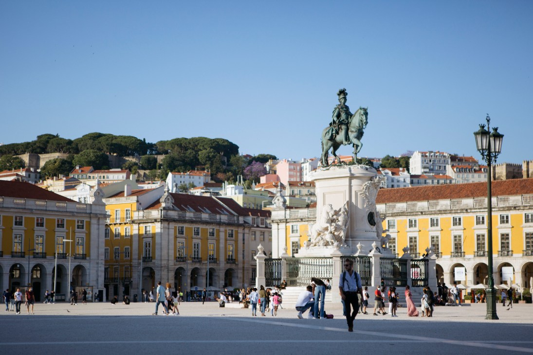 Terreiro do Paço square in Lisbon with equestrian statue, colorful historic buildings, and crowds of people under clear blue sky.