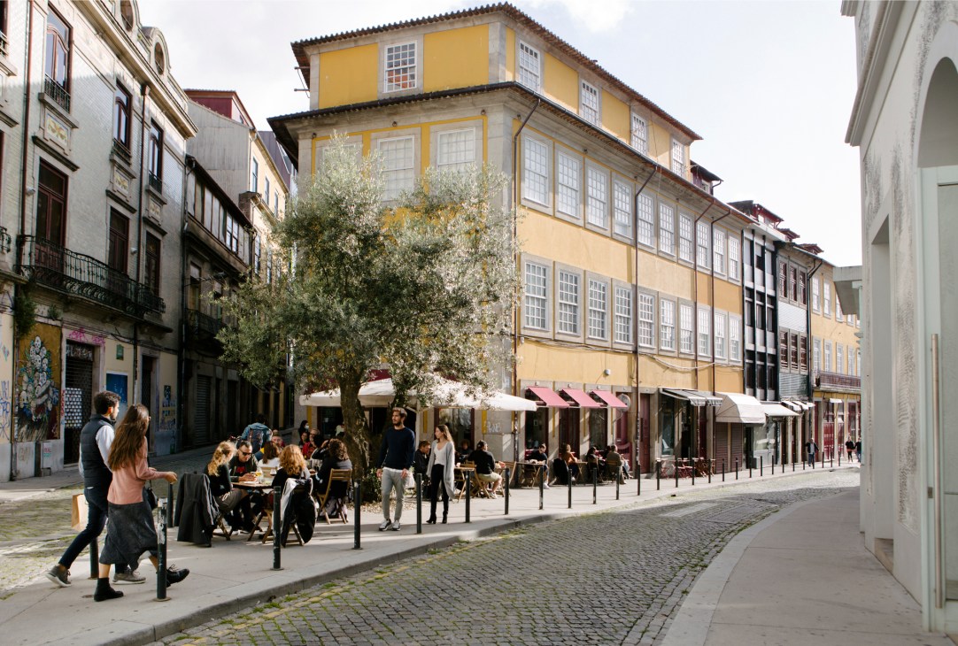 Porto's historic cobblestone street with yellow buildings, outdoor café seating, and people walking past shops.