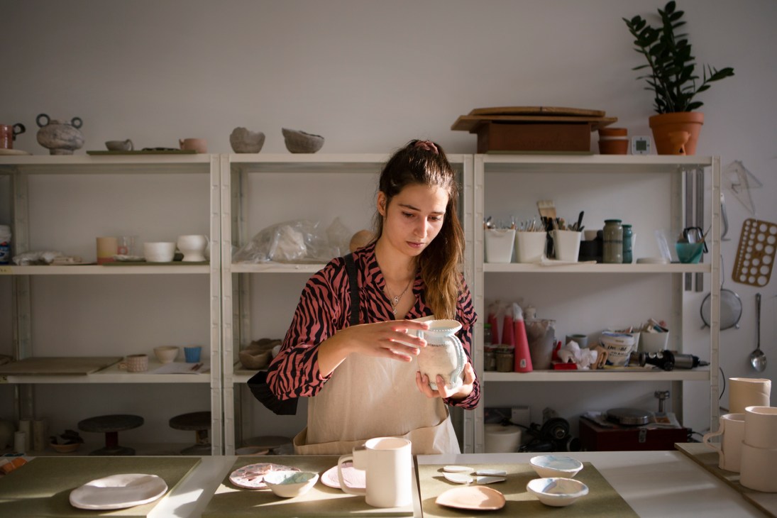 Woman in patterned shirt examining a large ceramic vessel in a studio with shelving displaying pottery and craft supplies.