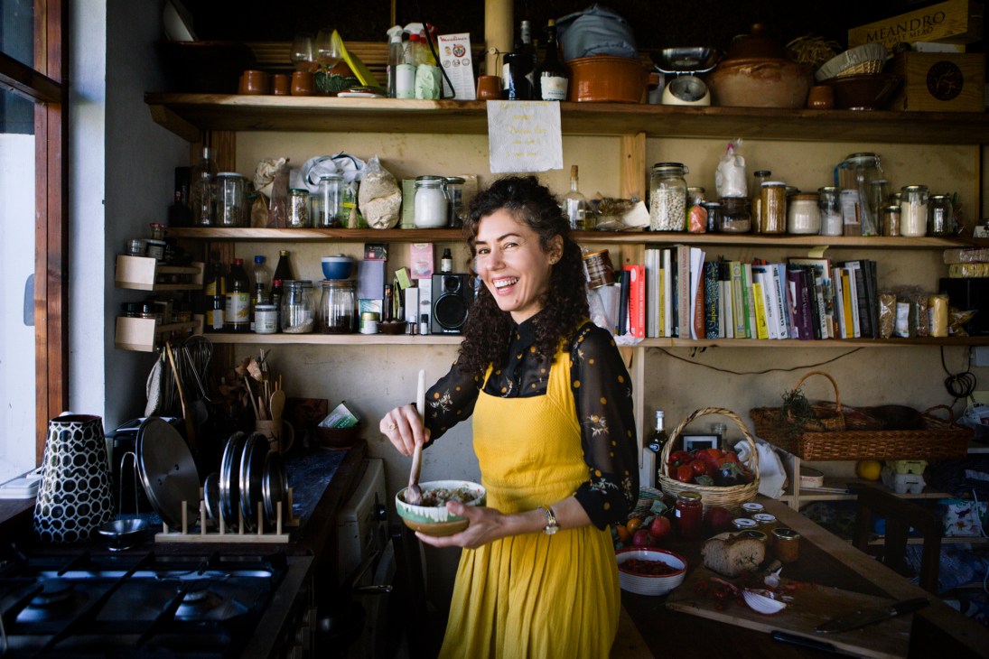 Woman in yellow apron smiling in a kitchen with shelves of ingredients and cookbooks.