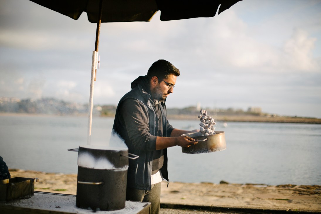 Man in dark jacket cooking oysters over fire on beach with water and landscape in background.
