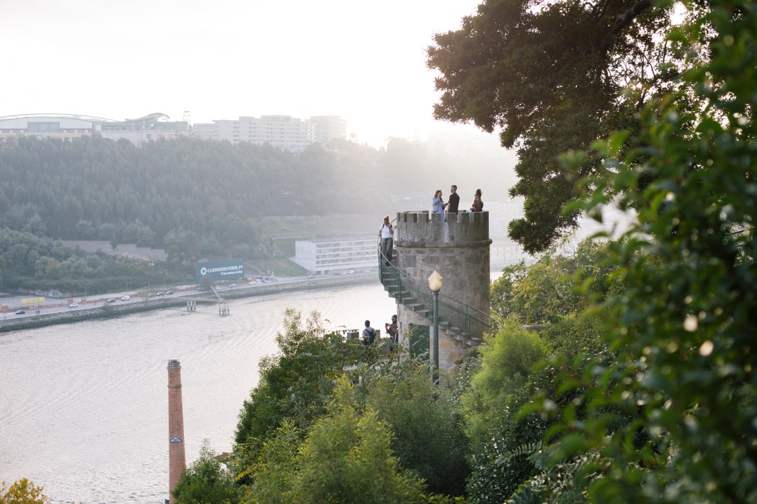 People standing on a stone tower overlooking a river and forested hillside with city buildings in the distance.