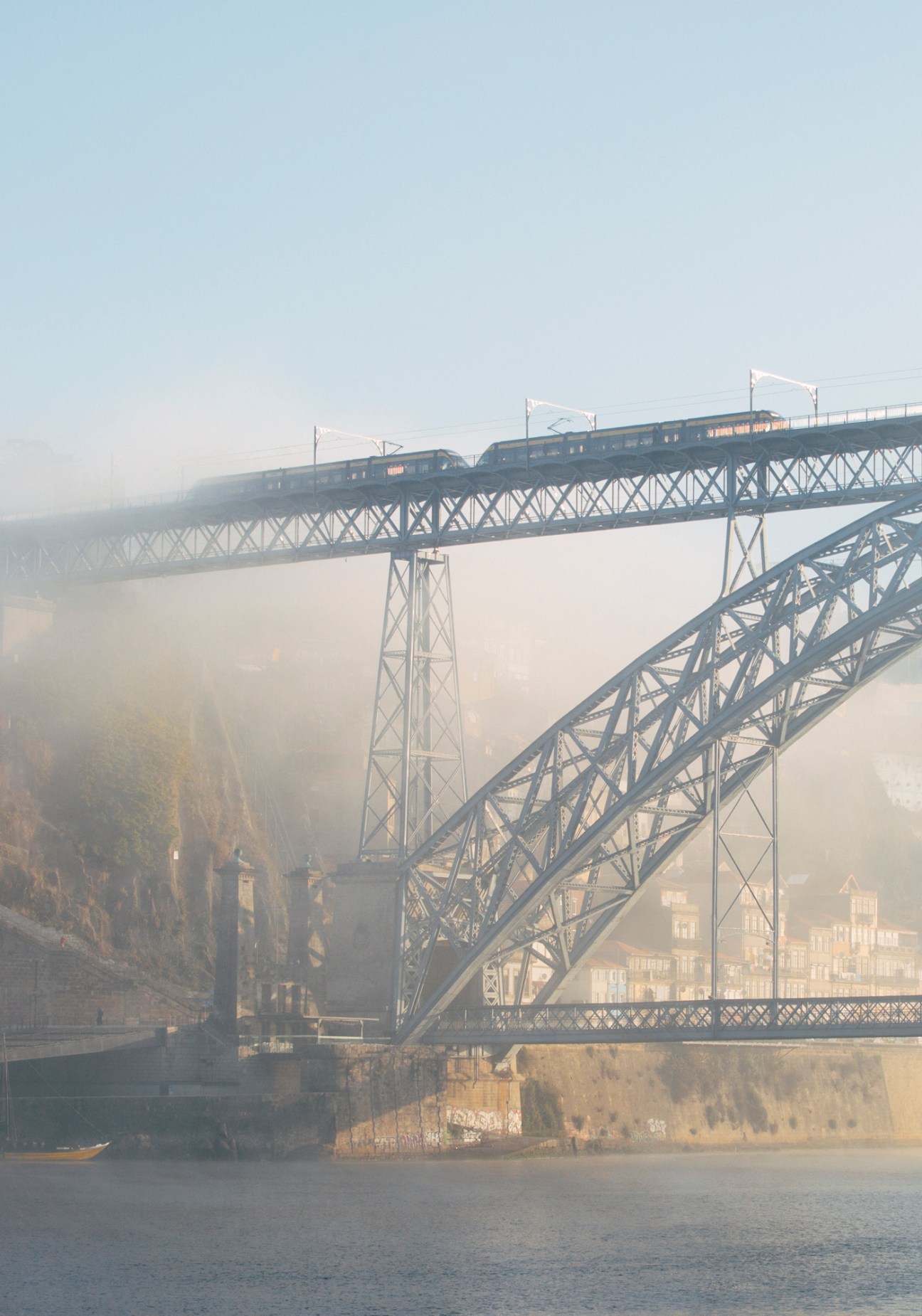A train crosses an ornate steel viaduct spanning a misty valley with forested hillsides below in Porto.