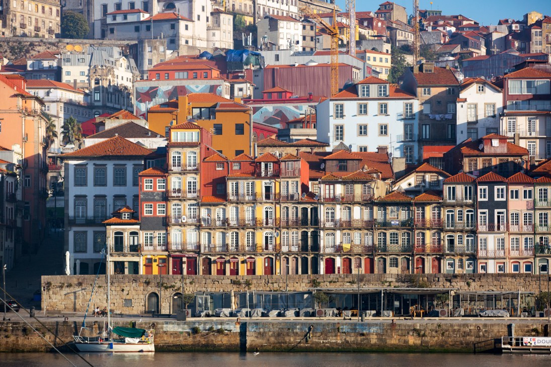 Colorful waterfront buildings with orange tile roofs line Porto's riverside, with boats moored below and terraced residential buildings climbing the hillside.