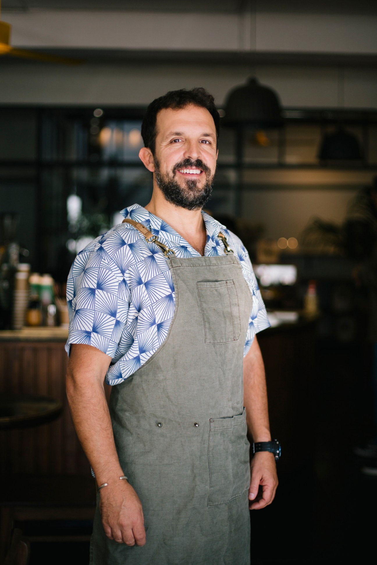 Man wearing blue patterned shirt and gray apron smiling in a restaurant kitchen