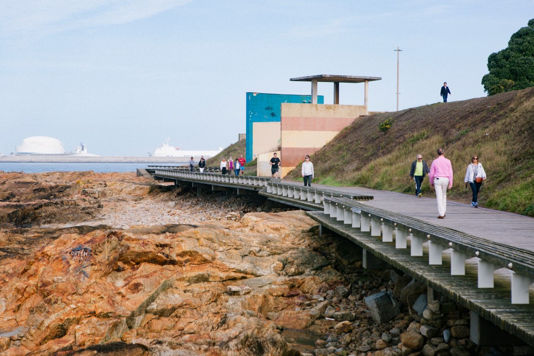 Wooden boardwalk with white railings extending along a rocky coastal landscape toward a colorful striped building with architectural features.