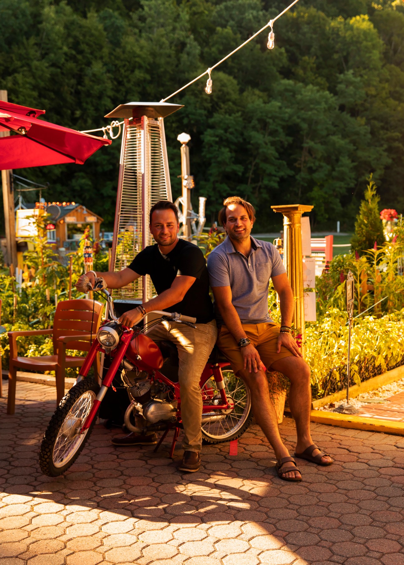 Two men posing with a red motorcycle on a patio area with outdoor dining furniture and string lights