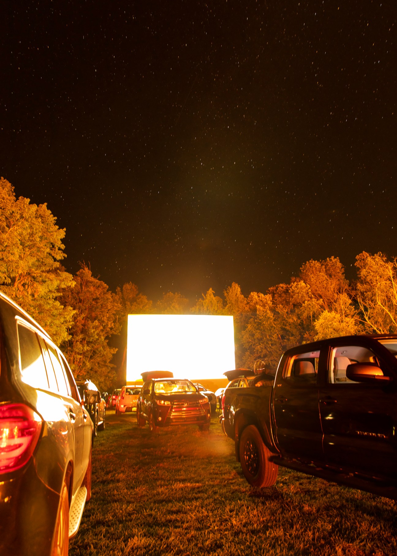 Cars parked at Four Brothers drive-in cinema at night with people sitting outside watching a movie under starry sky