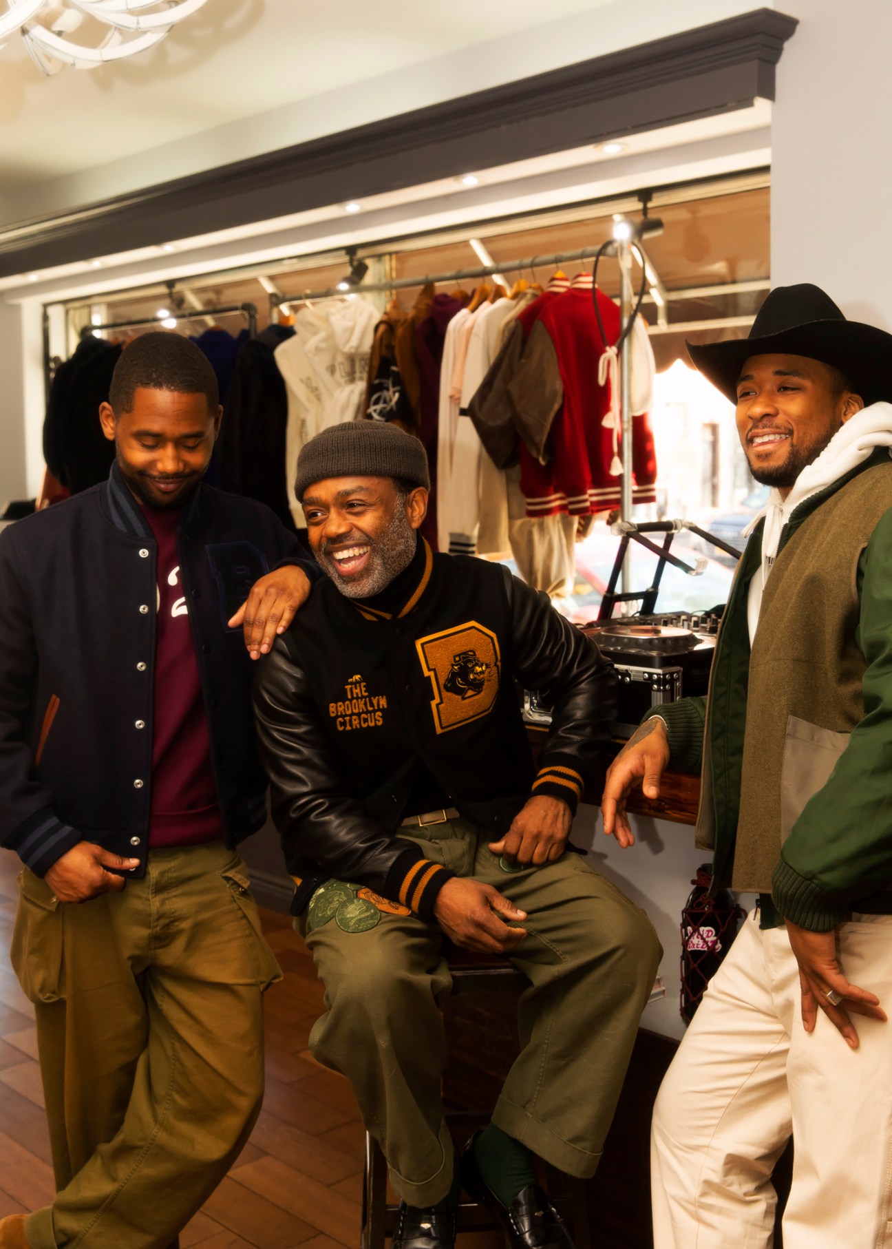 Three men in a clothing store with racks of menswear, one wearing a Brooklyn Circus leather jacket