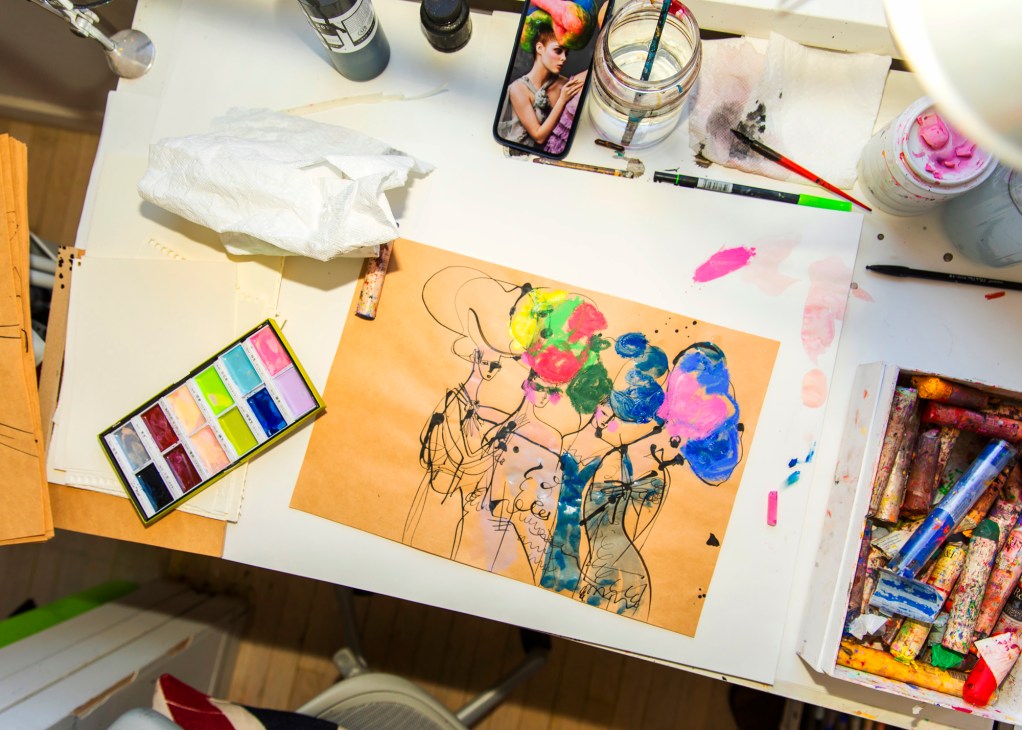 Artist's workspace with colorful fashion illustration on orange paper surrounded by paints, brushes and art supplies