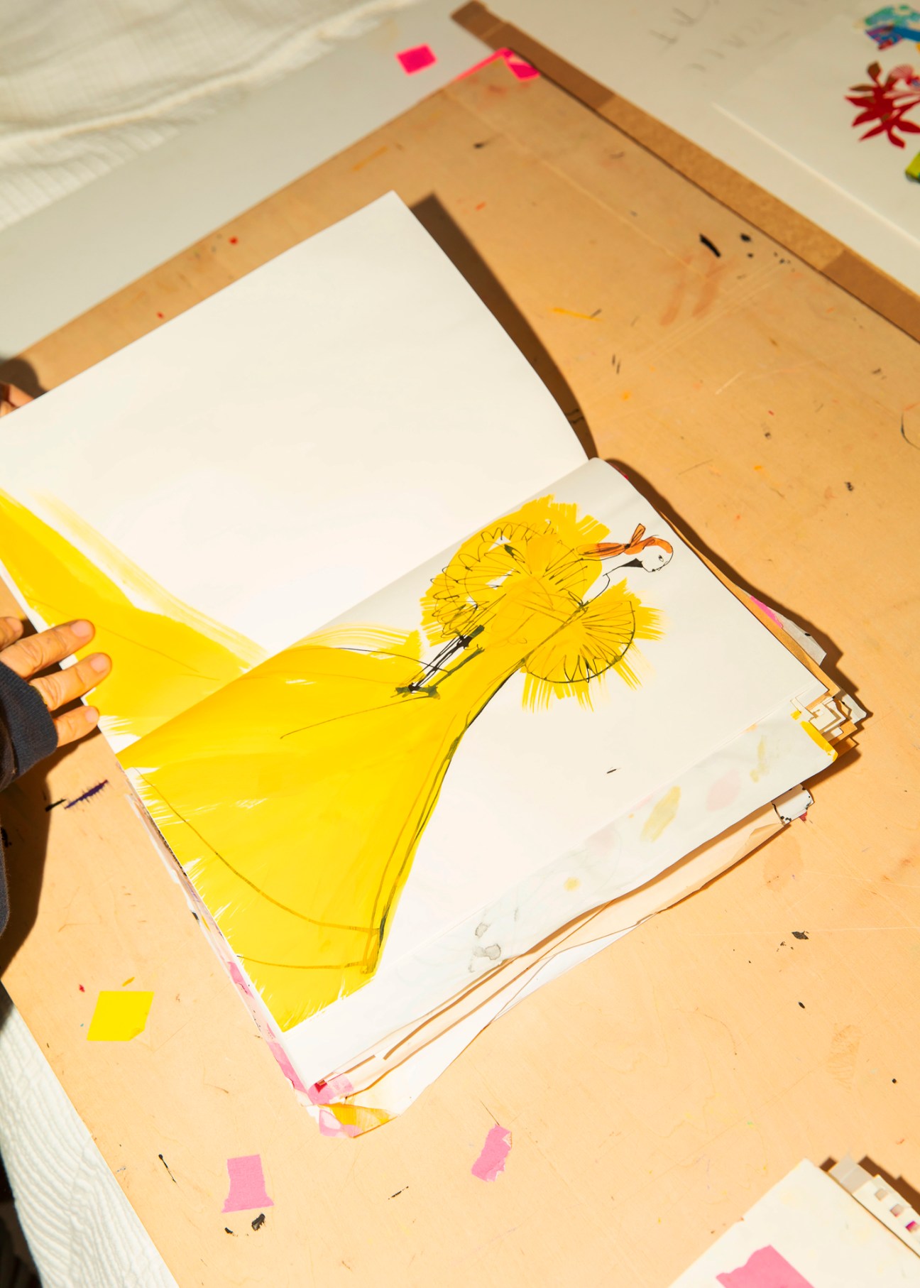 Hand holding yellow paper or fabric over an open sketchbook with fashion illustration on wooden drawing board