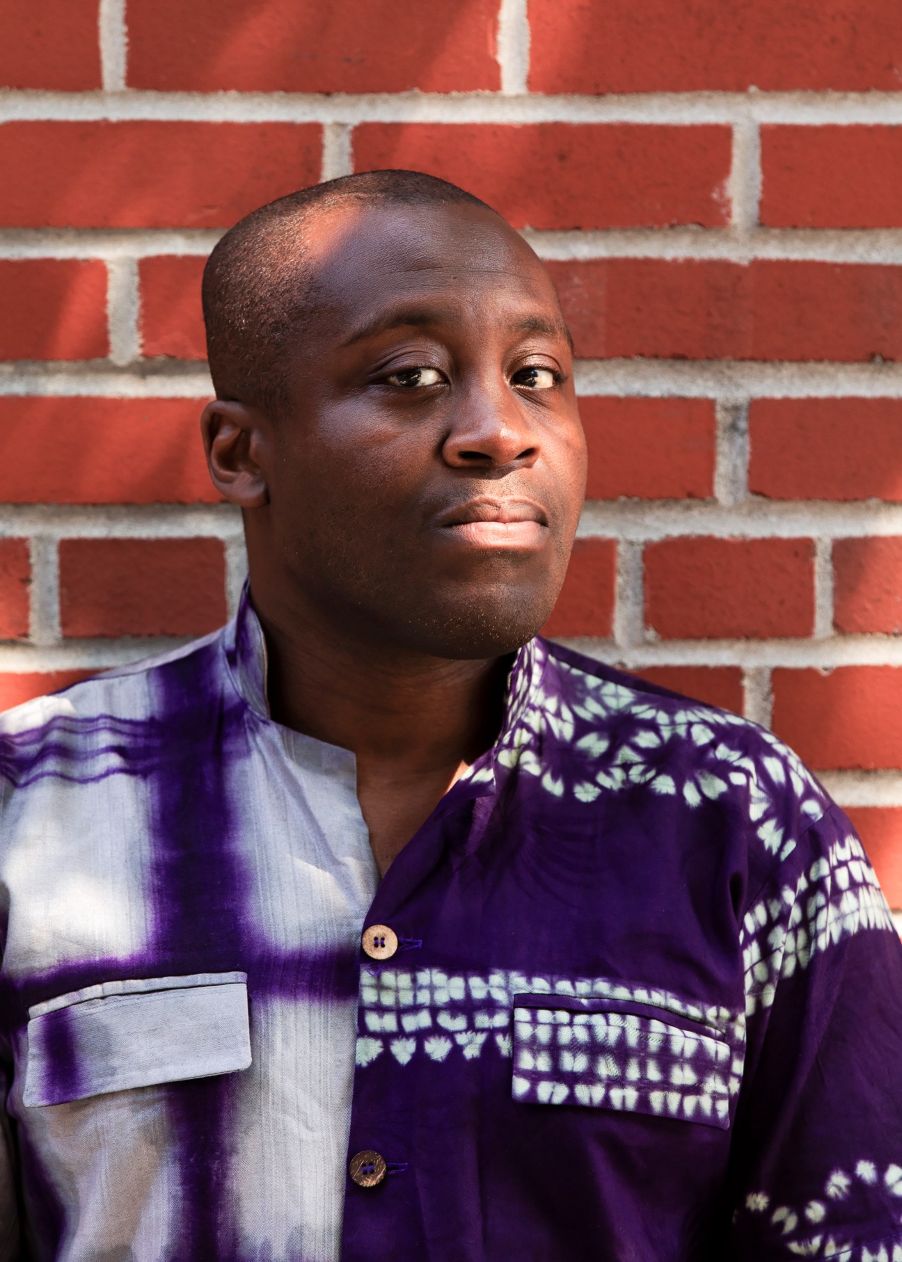 Larry Ossei-Mensah wearing a purple and white tie-dye shirt against a brick wall