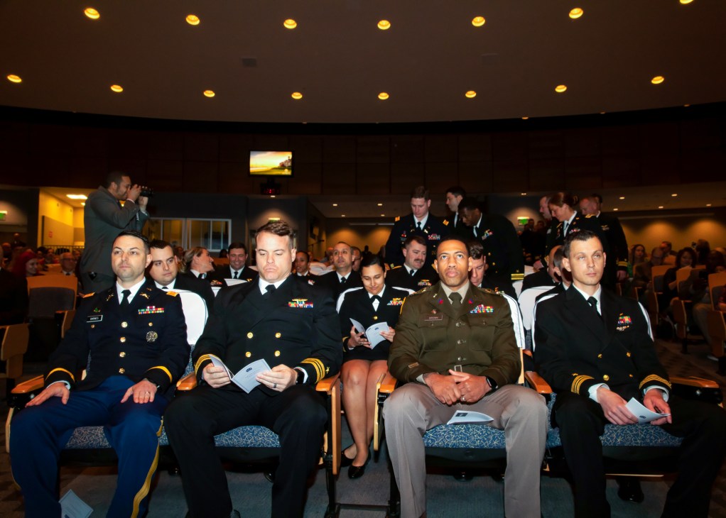 Military personnel in dress uniforms seated in auditorium chairs during graduation ceremony