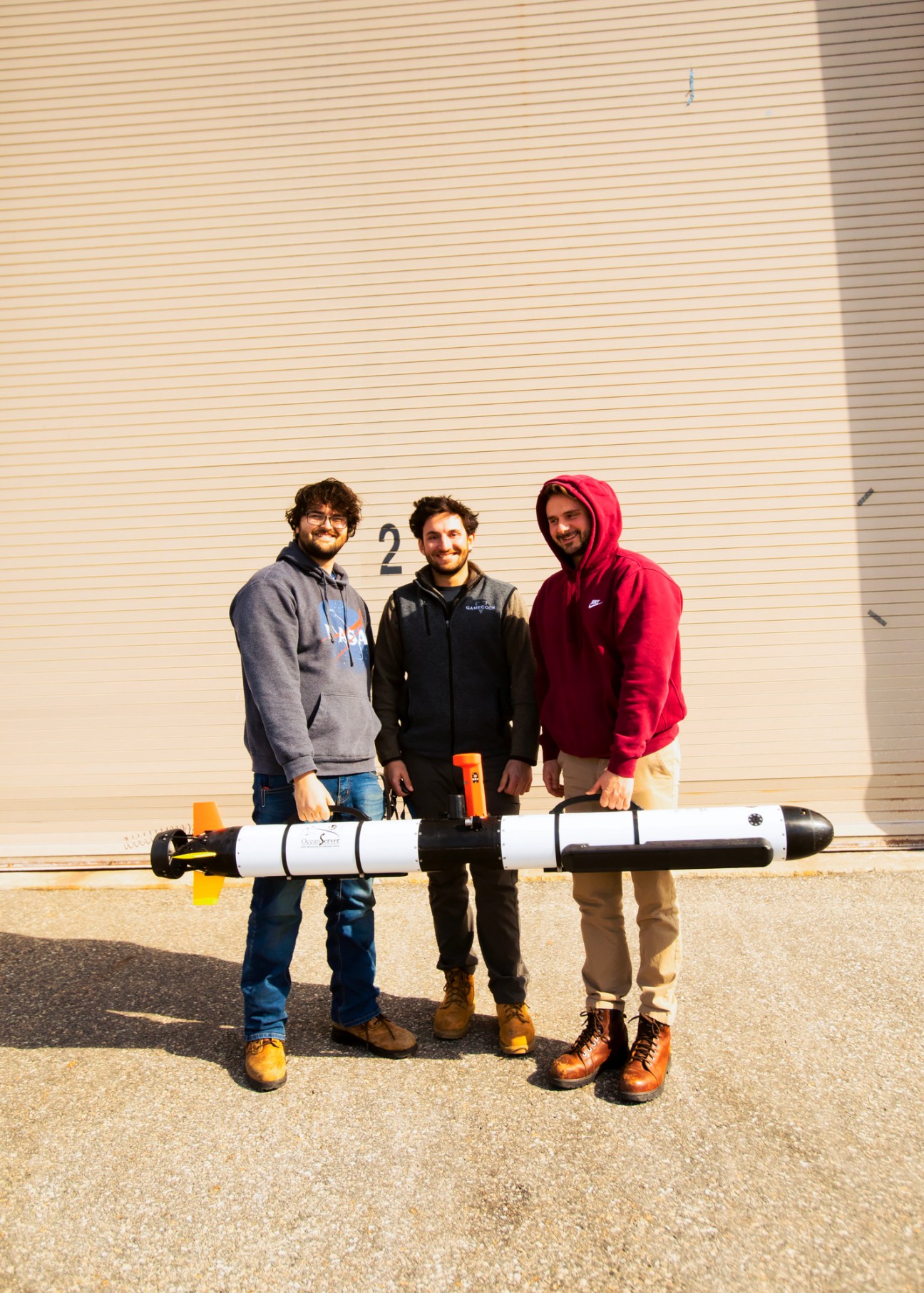Three men holding a white torpedo-shaped drone outside a beige building