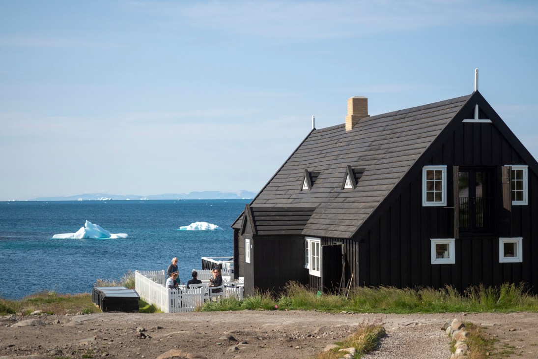 Dark wooden house with white-trimmed windows on coastal landscape with icebergs floating in blue water