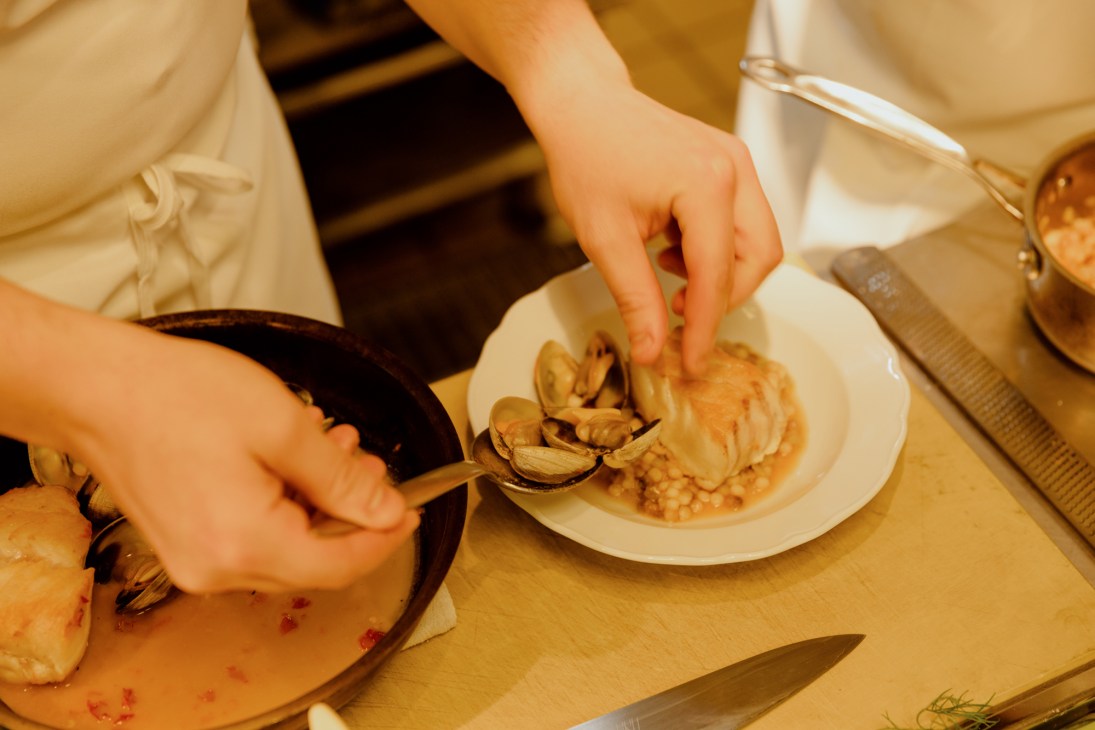 Hands plating seafood dish with clams and cod from black pan onto white plate