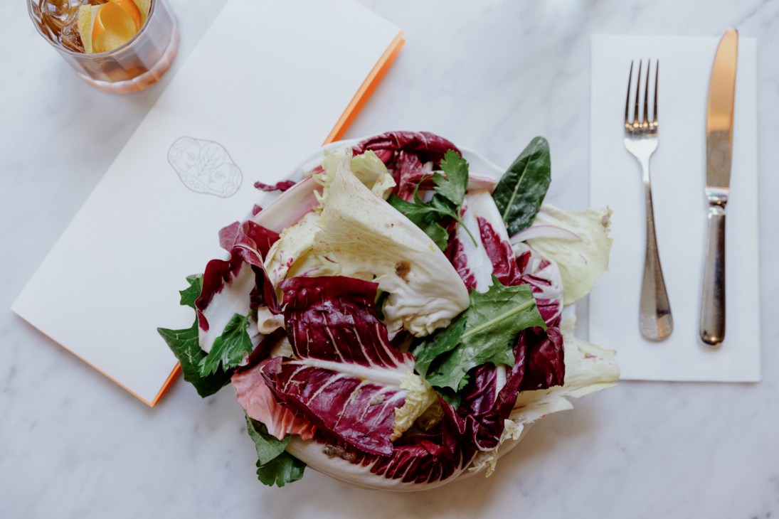 Bowl of mixed chicory and radicchio salad with white napkin, fork and knife, and glass on marble surface