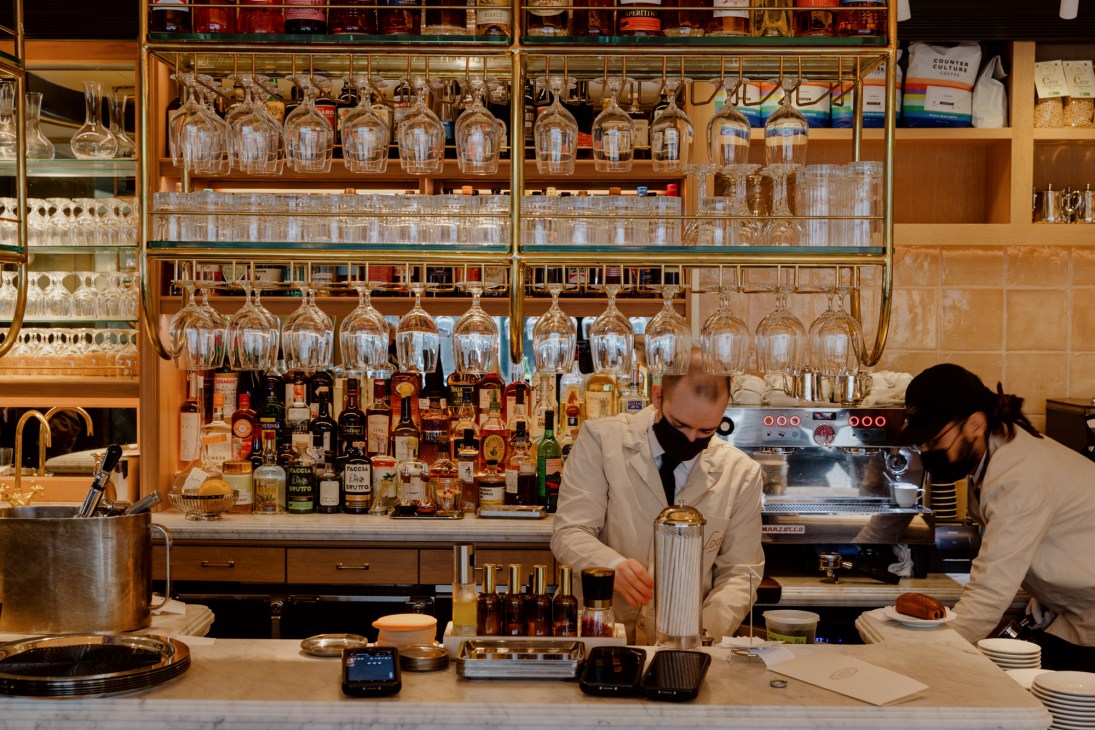Two bartenders in white uniforms working behind a well-stocked bar with hanging wine glasses and bottles