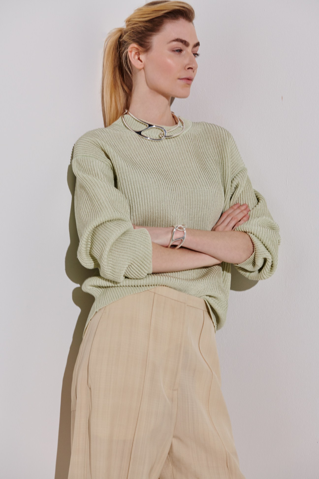 Woman in sage green ribbed jumper and beige trousers with chain necklace and bracelet