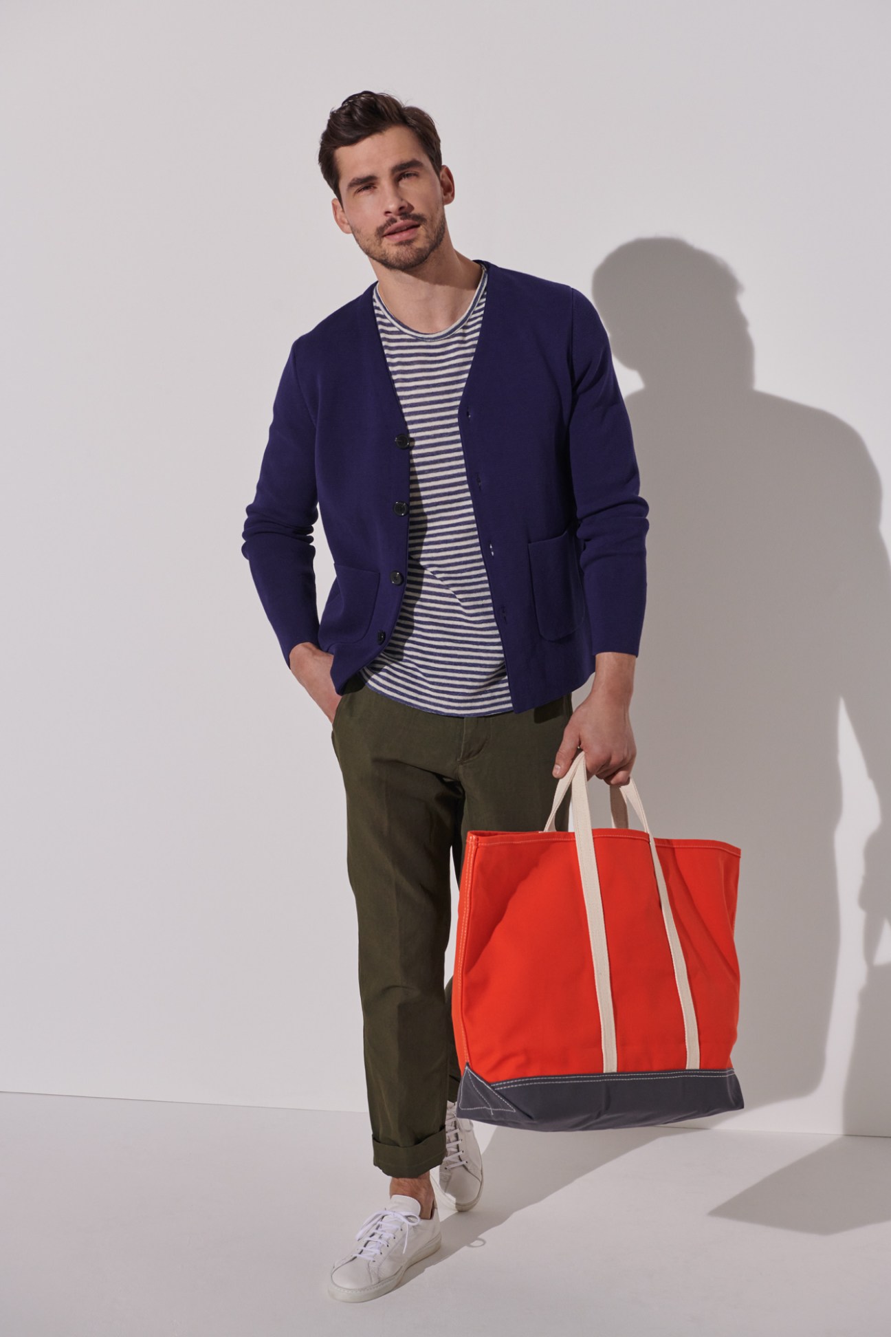 Man in navy cardigan, striped t-shirt, olive trousers and white trainers holding a red and navy canvas bag