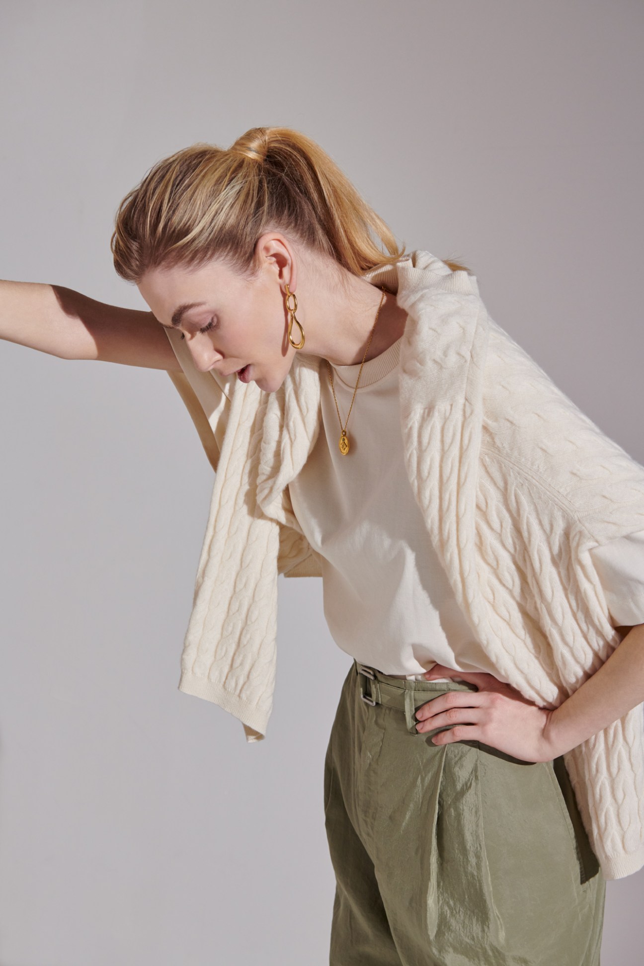 Woman in cream cable-knit sweater and olive green trousers with gold jewelry posed against neutral background