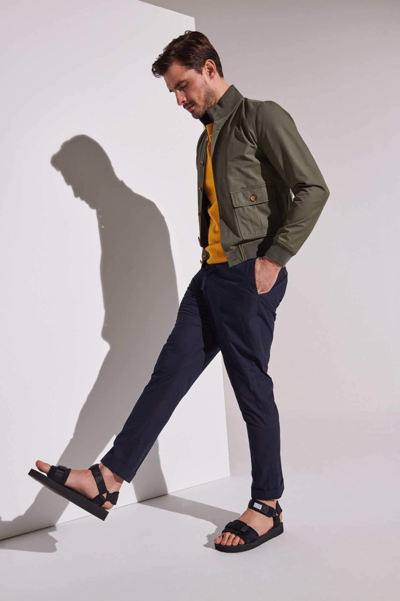 Man wearing olive green Valstar jacket over orange Canali jumper with navy A Kind of Guise trousers and Suicoke sandals