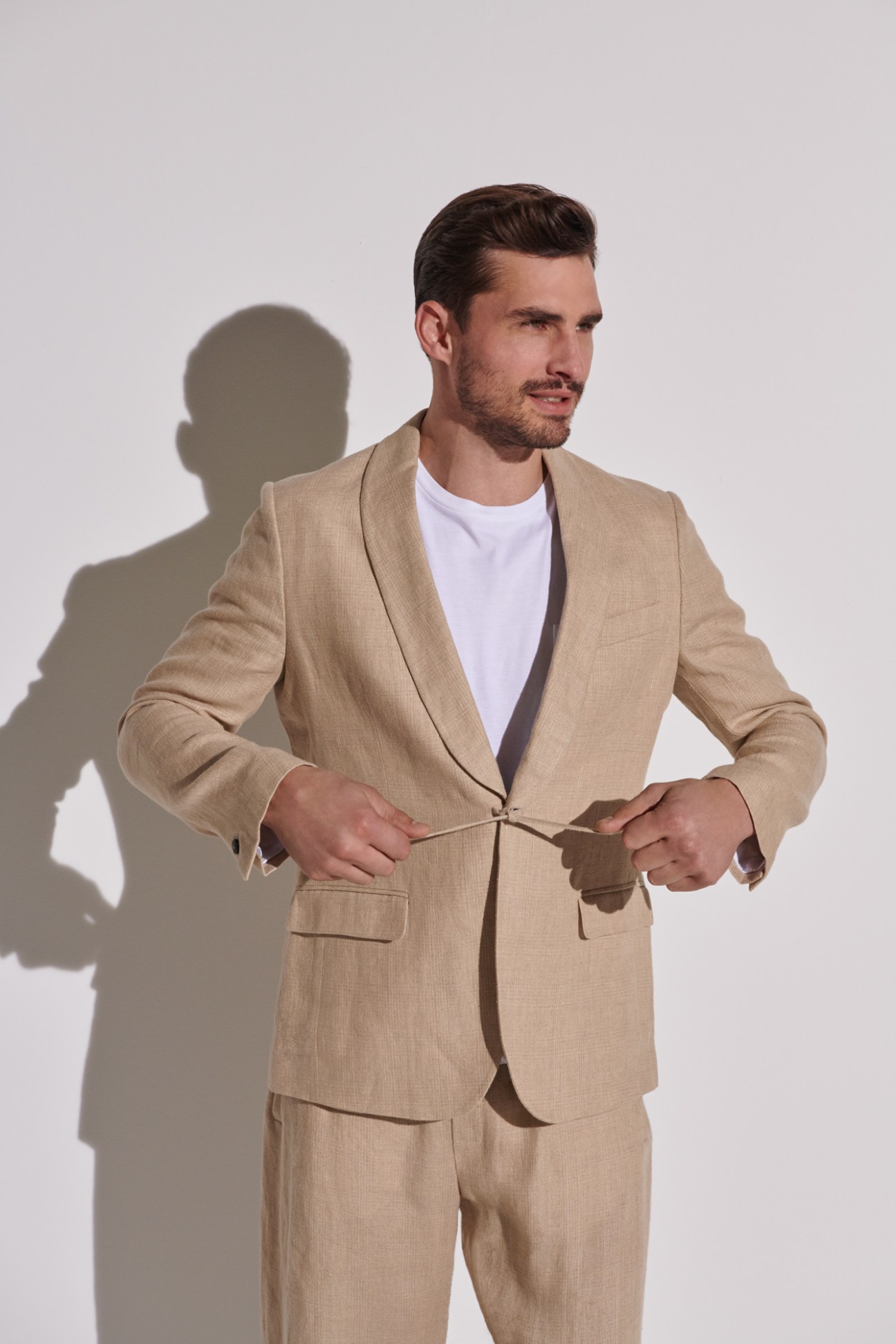 Man in beige linen suit and white t-shirt adjusting his jacket against white backdrop