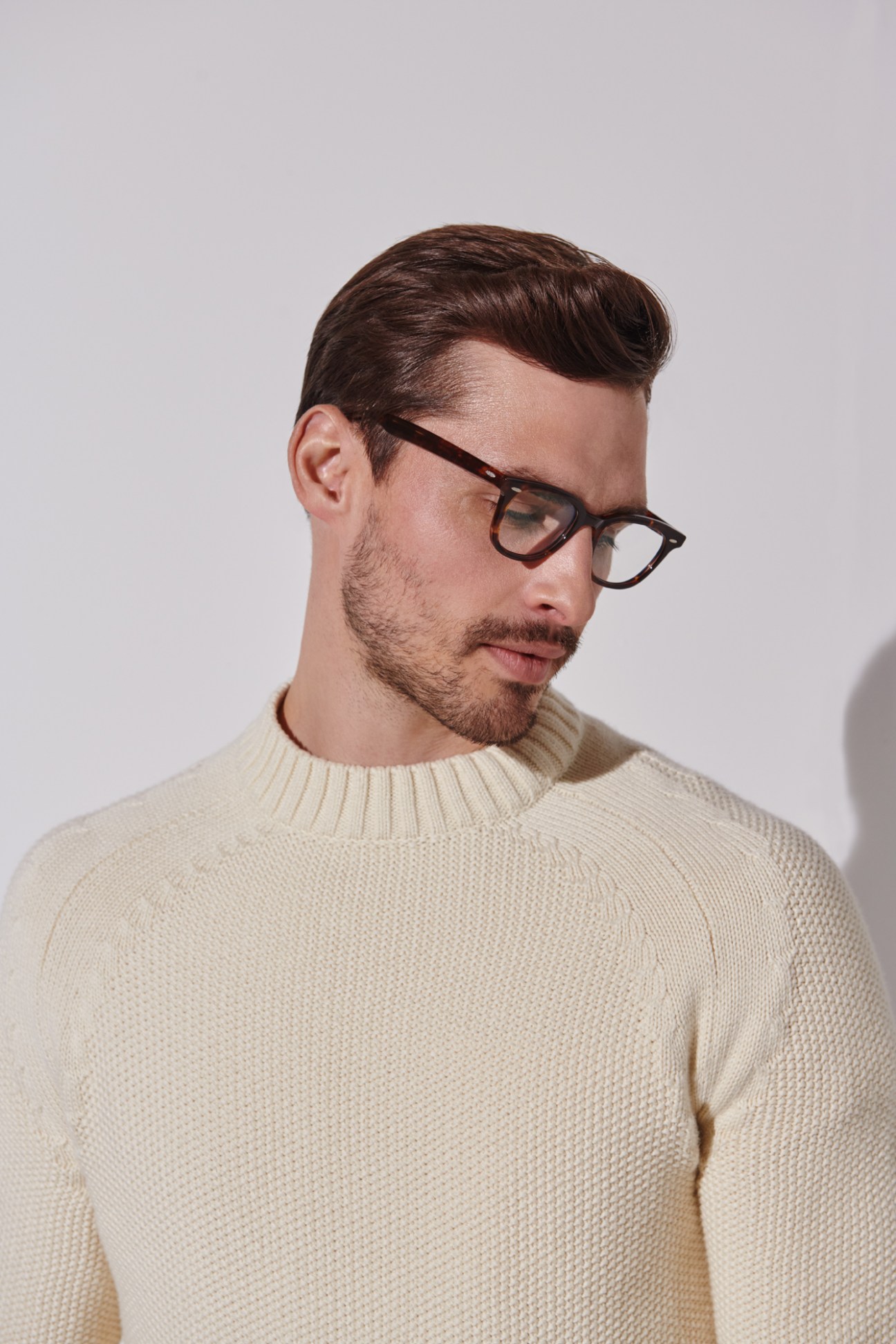Man in cream knit jumper and glasses looking to the side against neutral background