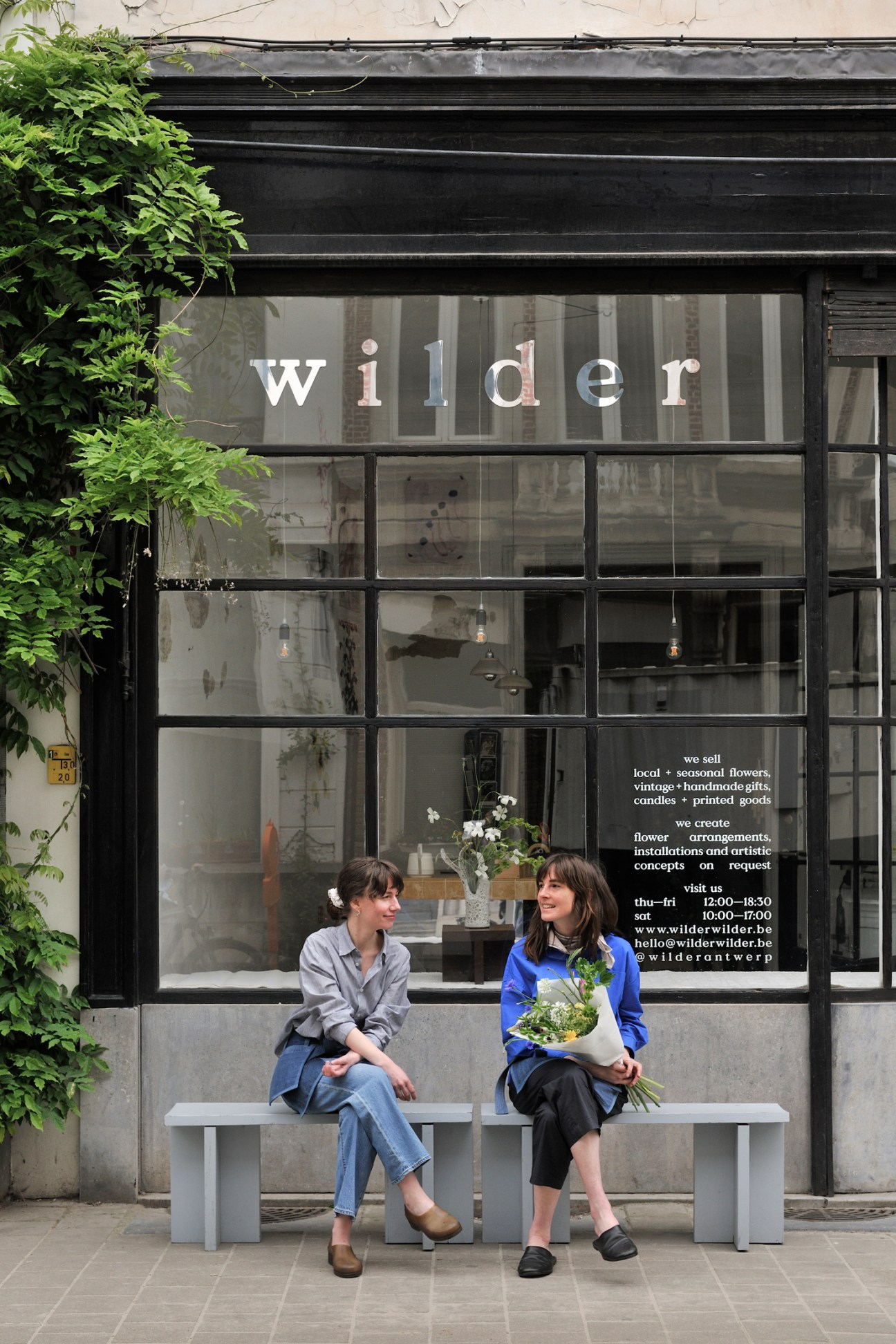 Two women sitting on a bench outside Wilder florist shop in Antwerp, one holding flowers