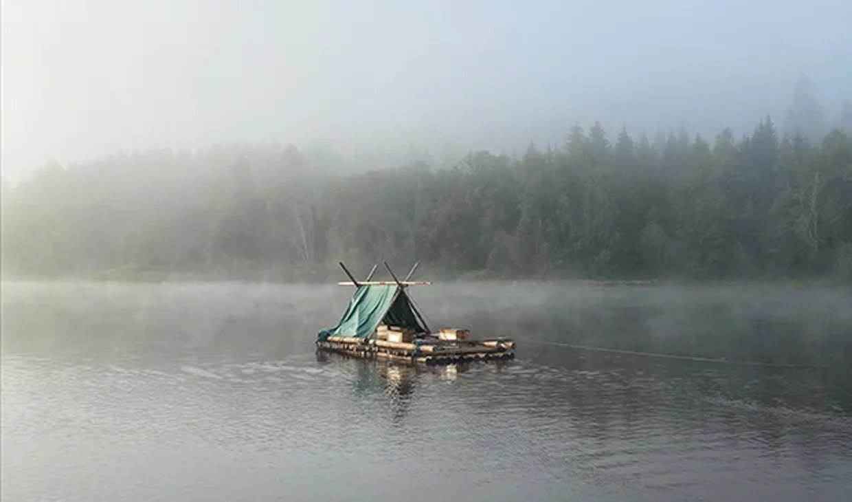 A couple sits in a tent on a homemade raft floating on a misty lake surrounded by forested shores.