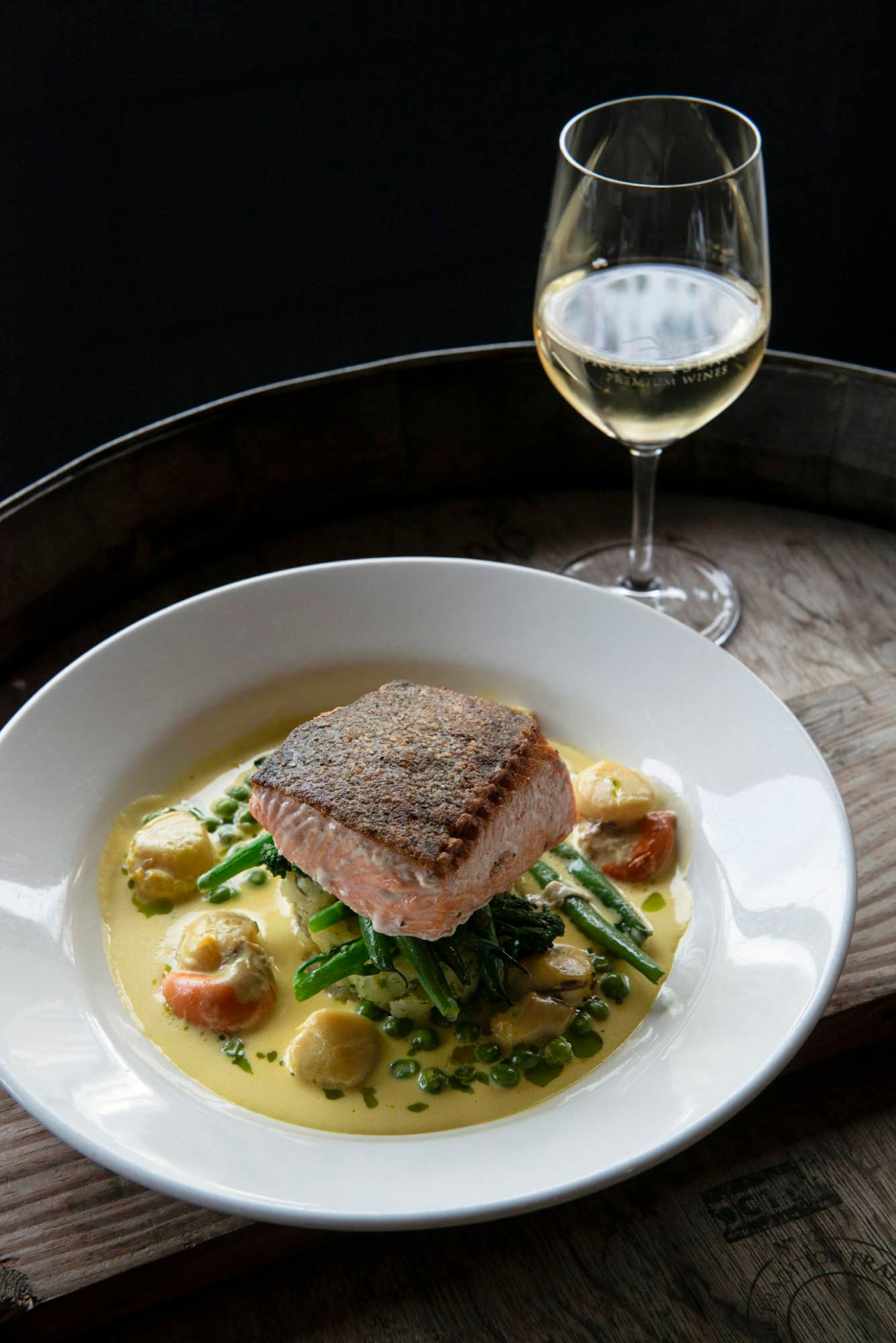 Seared fish fillet served on bed of greens with vegetables in creamy broth alongside glass of white wine