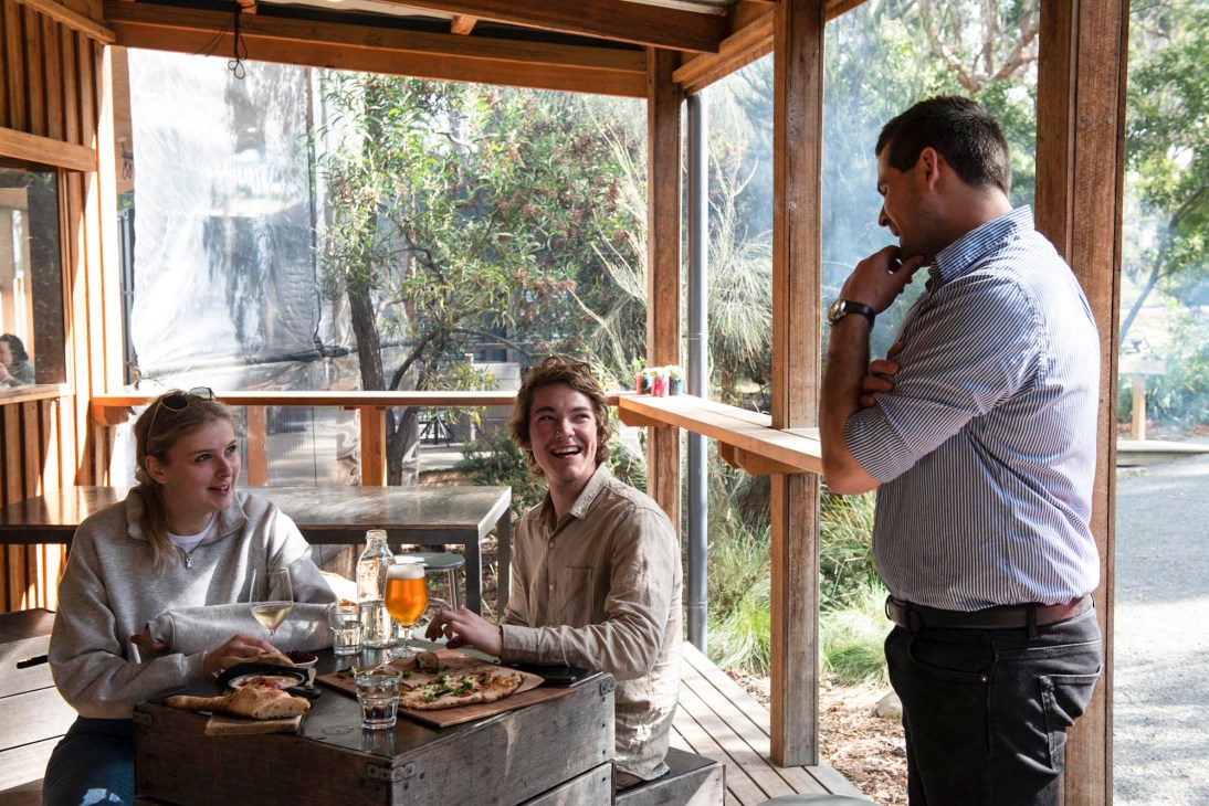 Two people dining outdoors at a wooden table while a man stands nearby at Bruny Island Cheese Company