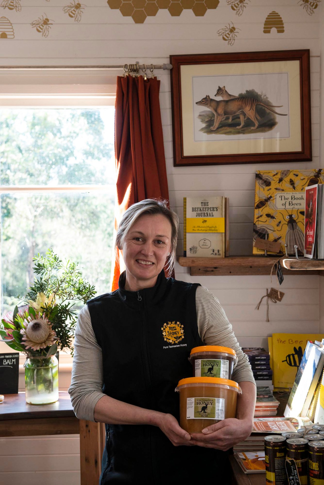 Woman in black apron holding two jars of honey in shop with bee-themed wallpaper and books
