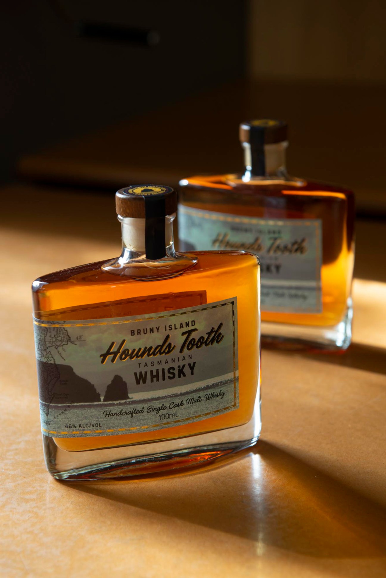 Two bottles of Houndstooth Distillery Tasmanian whisky with amber liquid and white labels on wooden surface