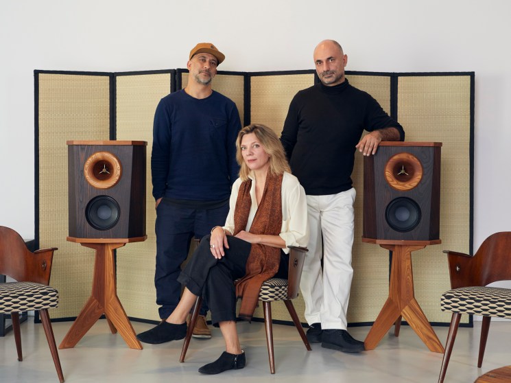 Three people posing with vintage wooden speakers in a studio setting.