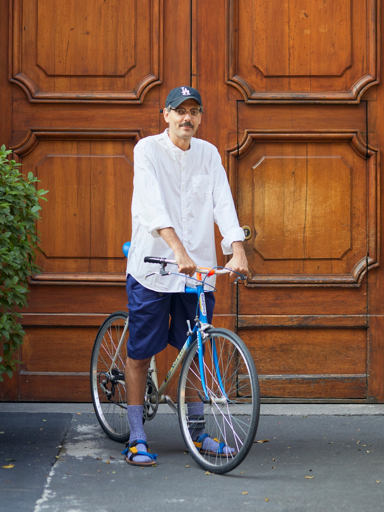 Man in white shirt and baseball cap standing with blue bicycle in front of ornate wooden doors
