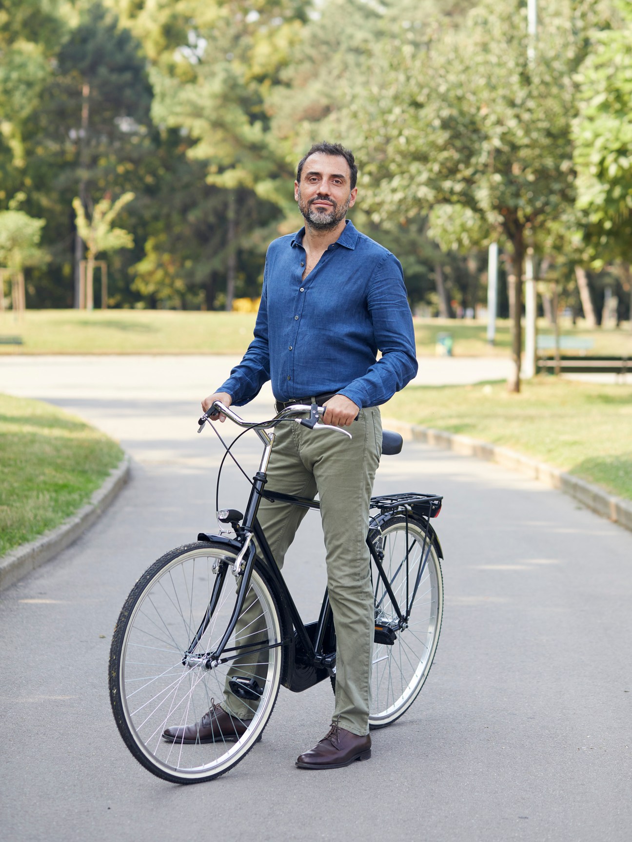 Man in blue shirt and khaki pants standing with black bicycle on tree-lined path in Milan