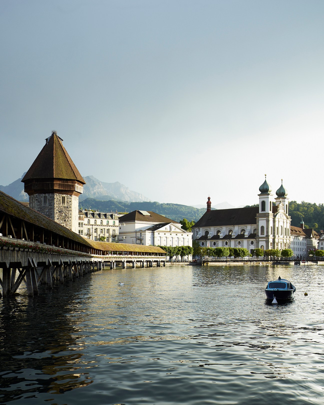 Lucerne's waterfront with historic buildings, chapel tower, and church spires reflected in the lake with Alpine mountains in the background.