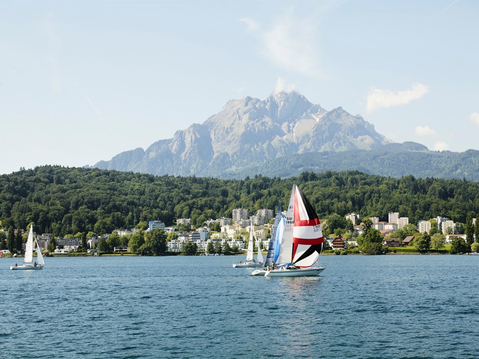 Sailboats with red and white sails on Lake Lucerne with snow-capped mountains in background.