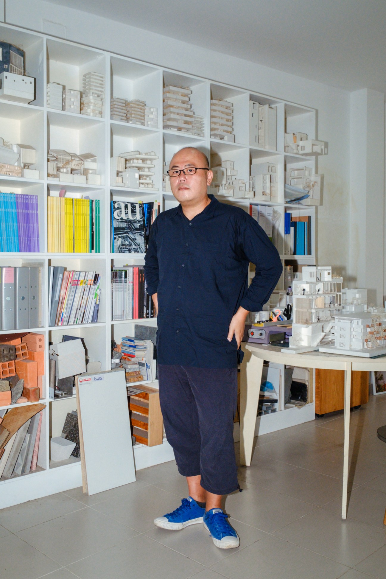 Japanese architect Sanuki Daisuke standing in his office in front of white shelving displaying architectural models and design books.