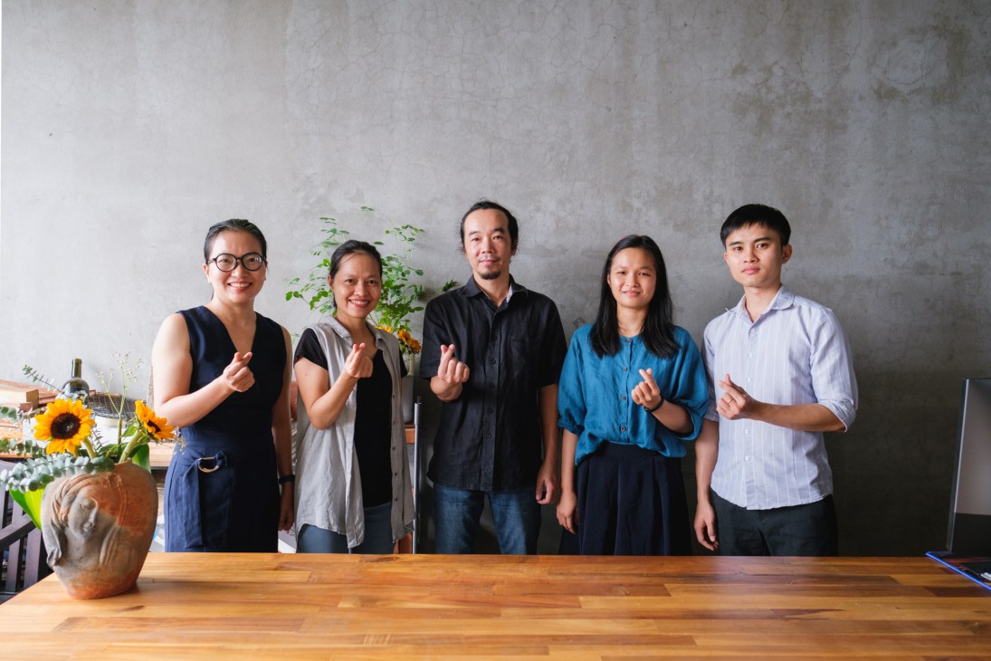 Five young architects stand together against a gray wall in a collaborative workspace with sunflowers and plants nearby.