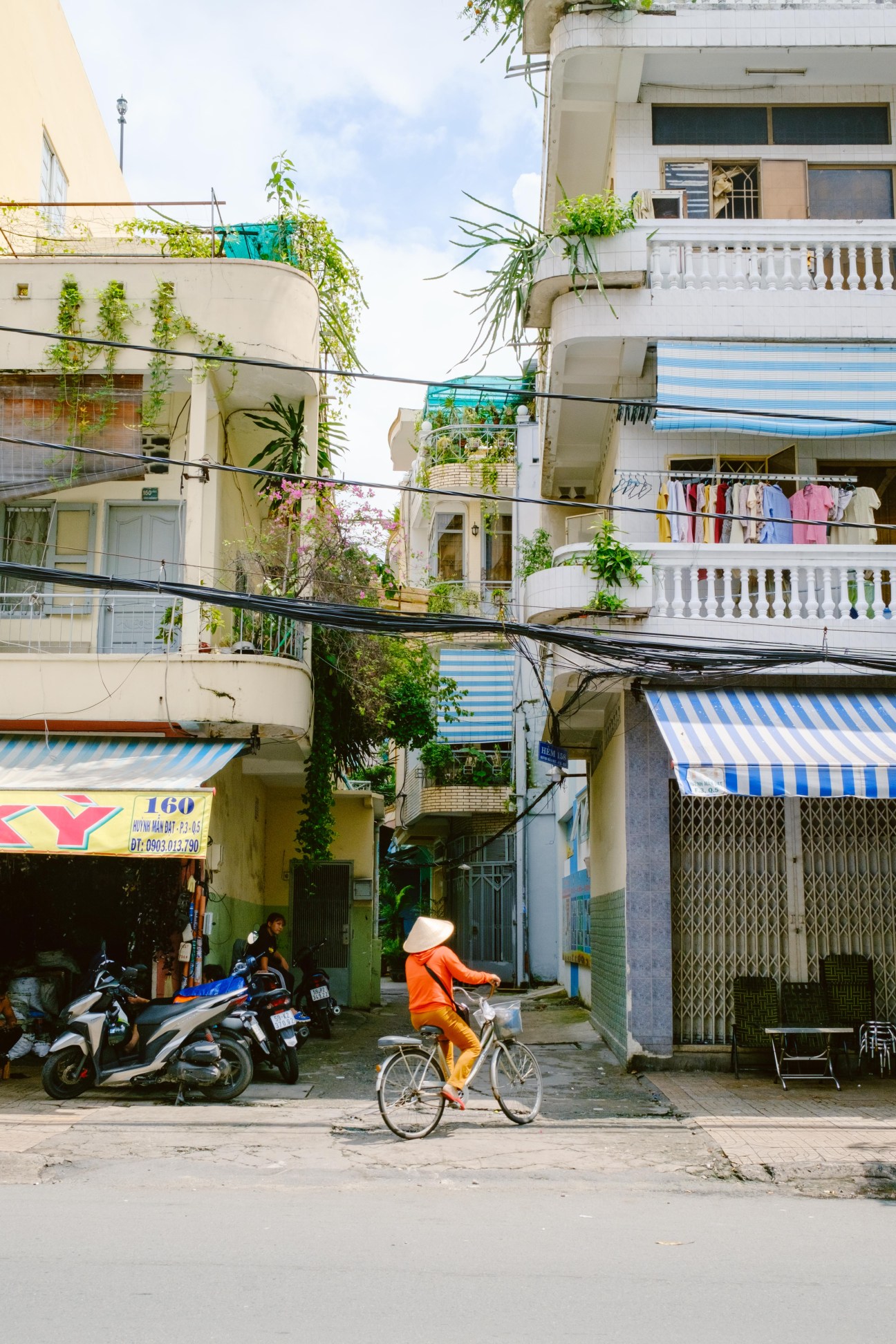A cyclist in an orange jacket riding through a narrow Ho Chi Minh City street lined with multi-story residential buildings and motorbikes parked below.
