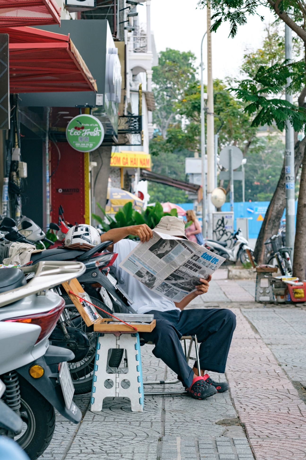 Man sitting on a scooter reading a newspaper on a Ho Chi Minh City street lined with shops and trees.
